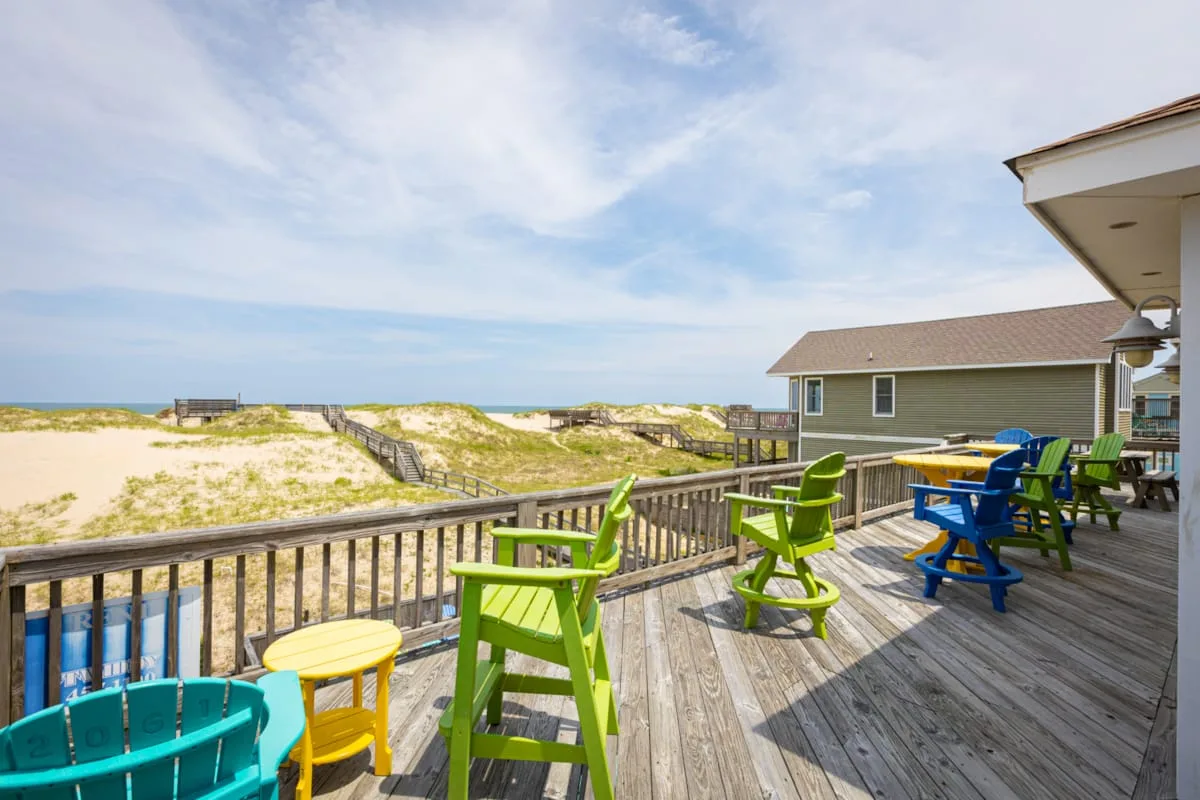 A wooden deck with colorful outdoor seating overlooks sandy dunes and the ocean.