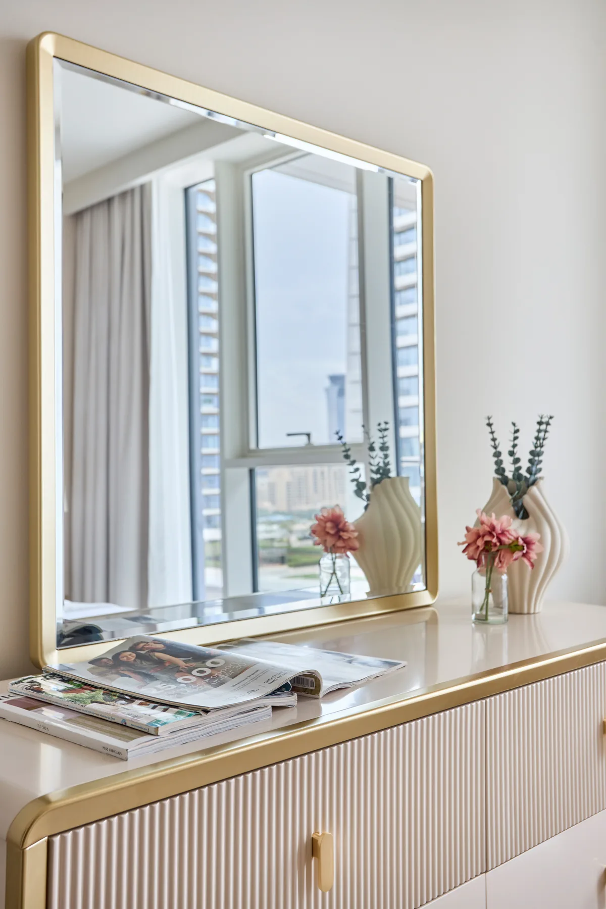 A bedroom dresser holds a large mirror, magazines, and small floral arrangements.