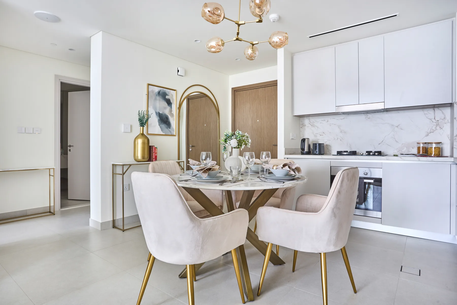 A bright dining area with a round table and pale upholstered seating beside a modern kitchen.