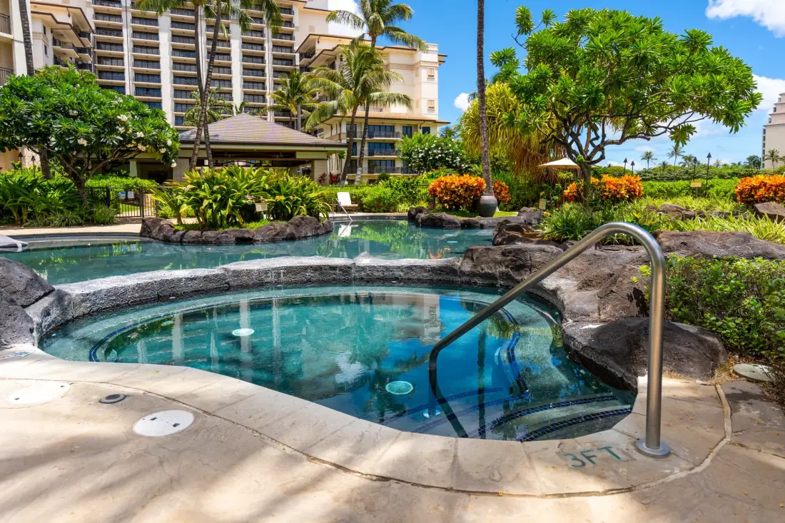 Una piscina al aire libre y un spa redondo están rodeados de vegetación tropical junto a un hotel.