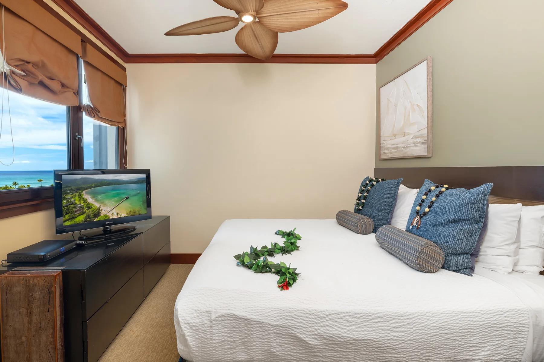 Un dormitorio con una cama bien tendida, cojines azules y un televisor junto a la ventana.