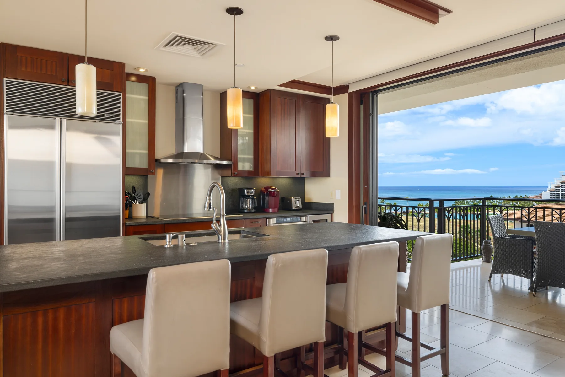 A kitchen with a long island and seating opens onto a balcony overlooking the ocean.