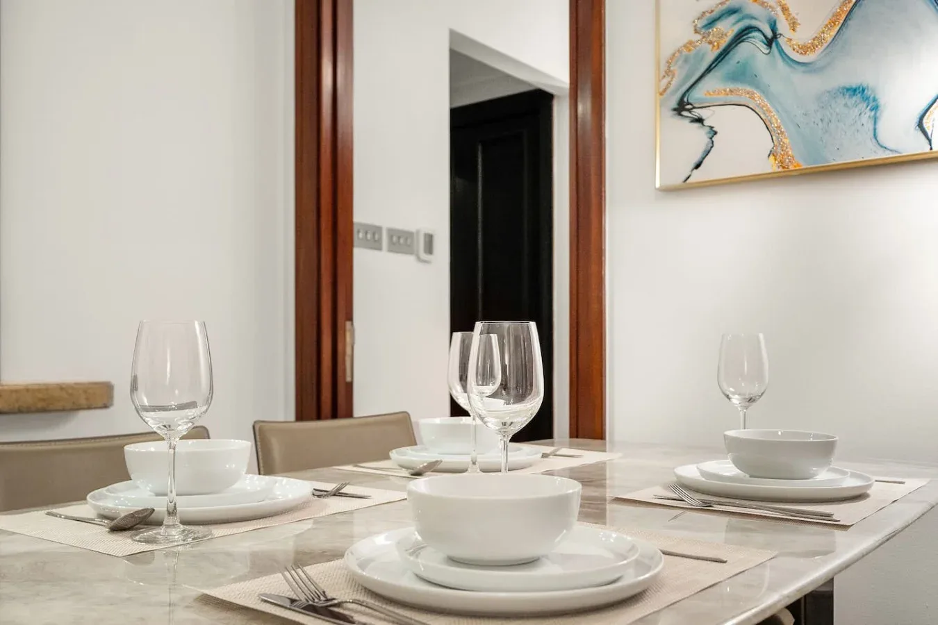 Una mesa de comedor está puesta con platos, cuencos y copas bajo un cuadro abstracto.