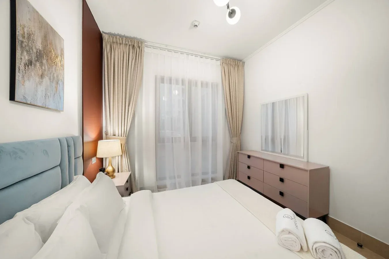 Un dormitorio con una cama, una lámpara de noche y una cómoda junto a una ventana con cortinas.