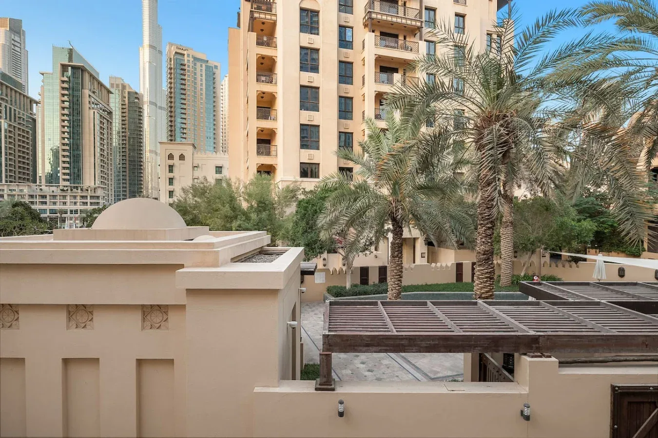 Un patio urbano con palmeras, una pérgola sombreada y edificios altos alrededor.