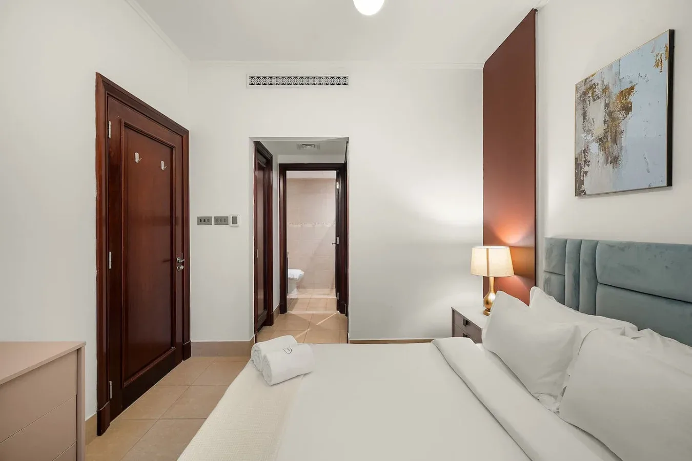 Un dormitorio con la cama tendida, una lámpara de noche y una puerta hacia el baño.