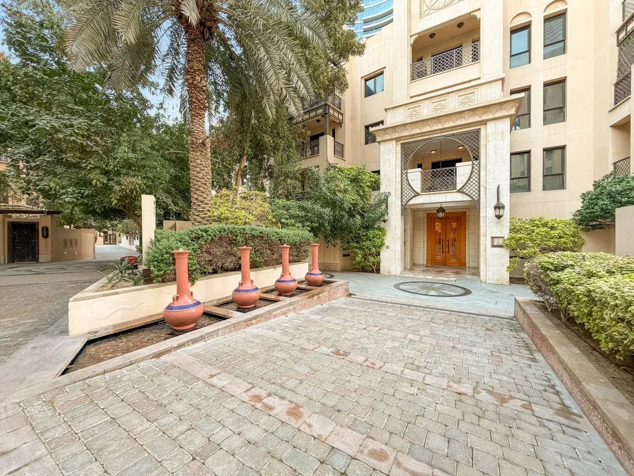 Un patio pavimentado con palmeras y jardineras está frente a la entrada de un edificio.