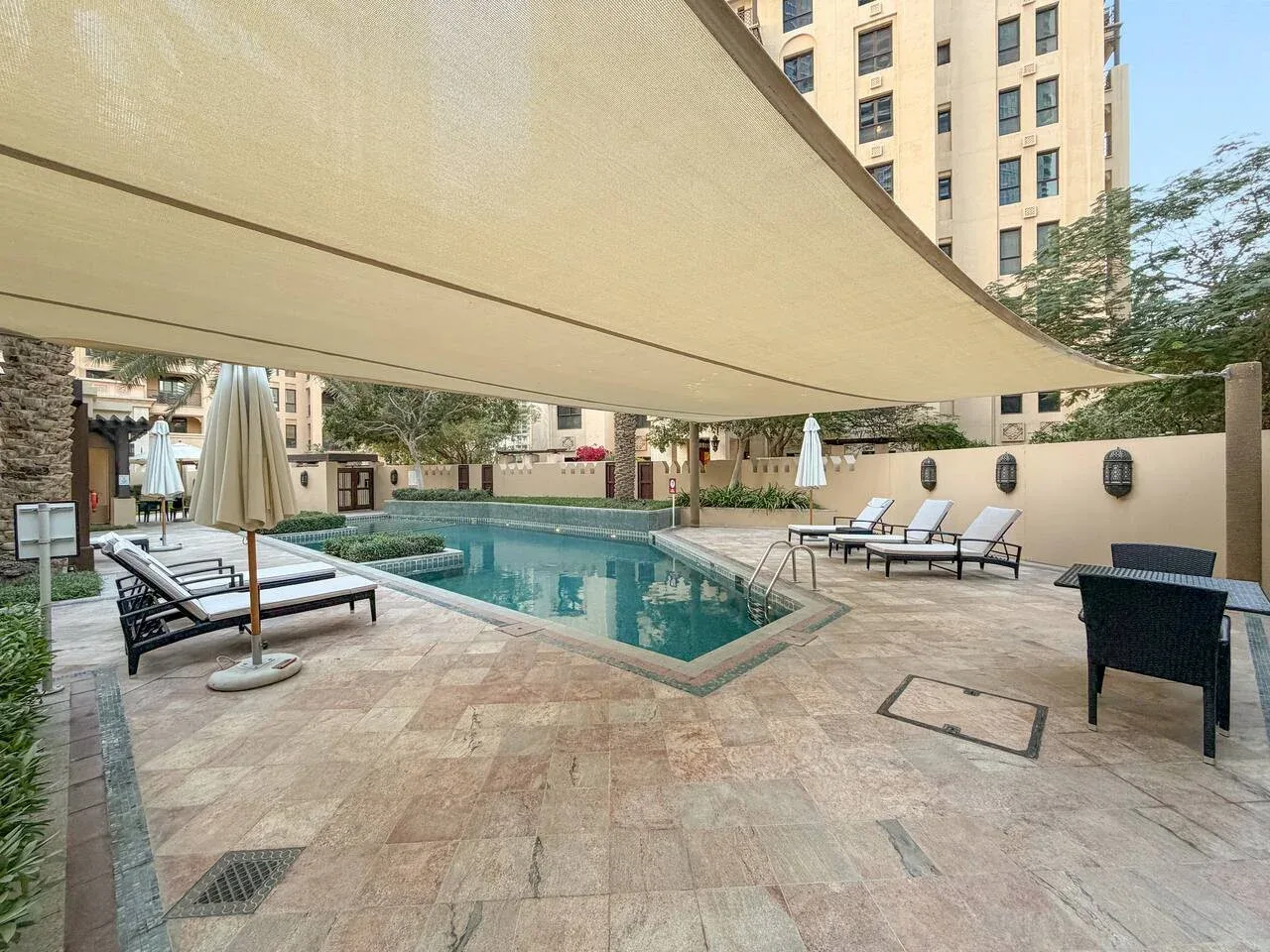 Zona de piscina exterior a la sombra, con tumbonas y edificios alrededor.