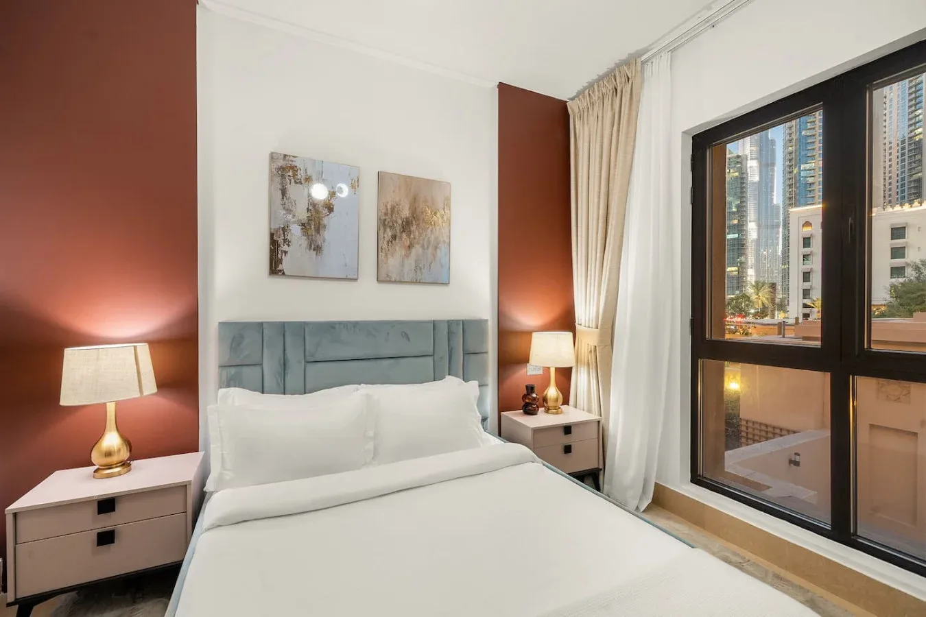 Un dormitorio con una cama bien arreglada y una ventana grande con vistas a edificios cercanos.