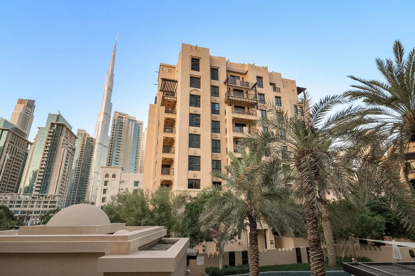 Un edificio residencial urbano se alza entre palmeras, con el Burj Khalifa visible a lo lejos.