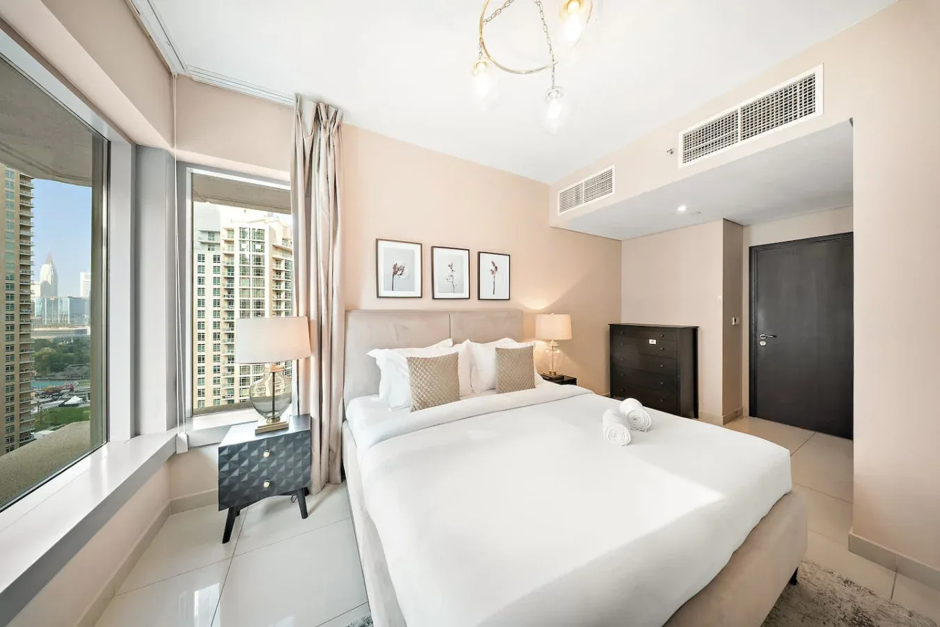 Un dormitorio luminoso con una cama grande junto a ventanas altas que dan a edificios cercanos.