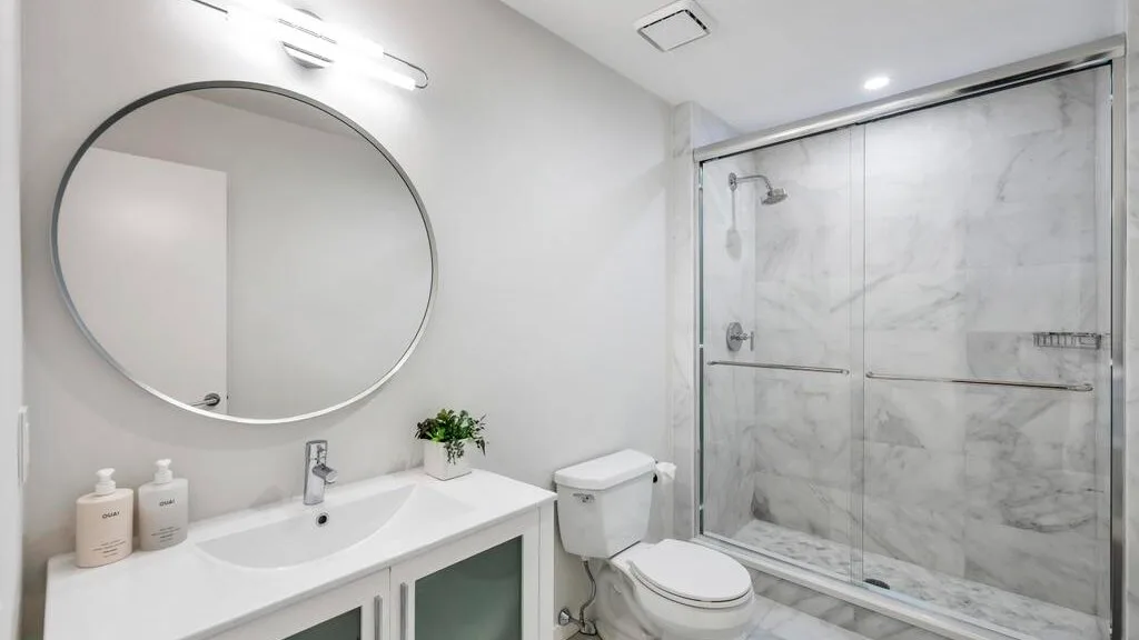 A bright bathroom with a sink, round mirror, and glass shower.
