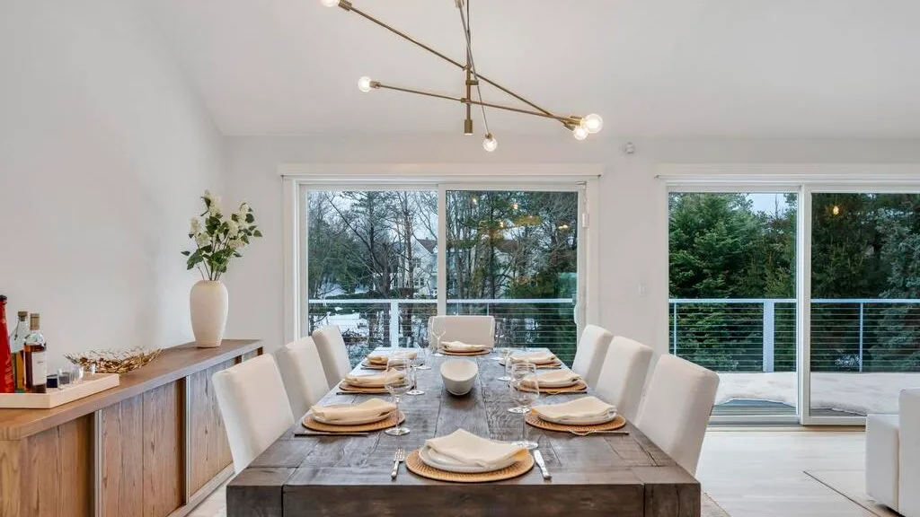 A dining area with a long table set beside large windows overlooking greenery.
