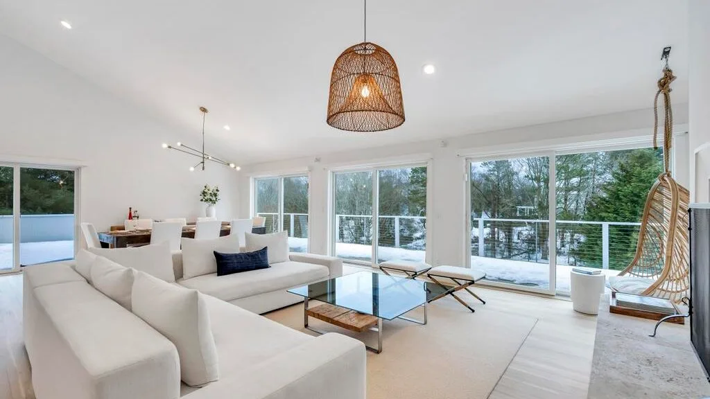 A bright living room with seating, a glass coffee table, and large windows.