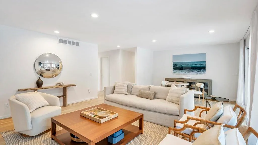 A bright living room with light seating, a wooden coffee table, and a coastal wall print.