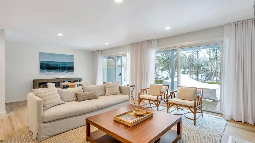 A bright living room with seating and a wooden coffee table beside large windows.