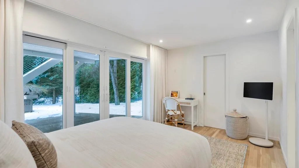 A bright bedroom with a bed beside large windows overlooking trees.