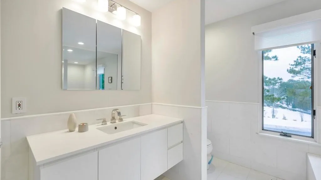 A bright bathroom with a long white vanity, mirrored cabinets, and a window.