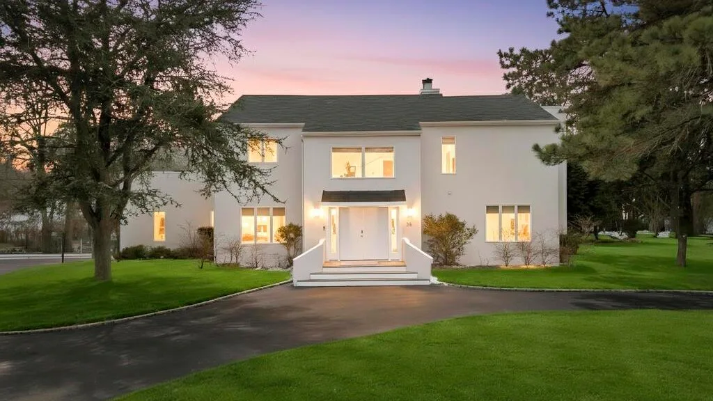 A white house with lit windows stands beyond a lawn and driveway at dusk.