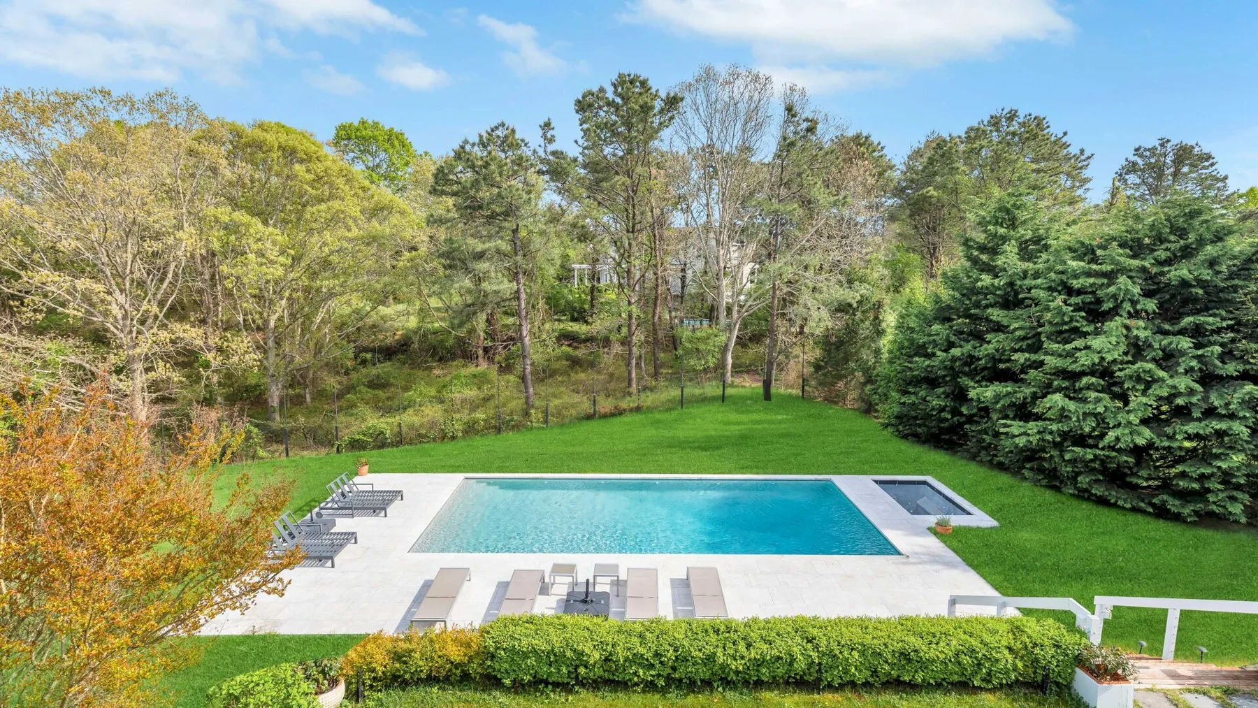 An outdoor swimming pool sits on a paved patio beside a green lawn and trees.