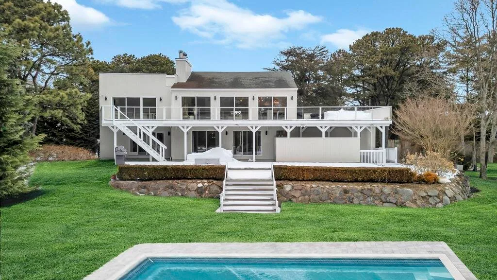 A white house overlooks a swimming pool, with a broad lawn and trees around it.