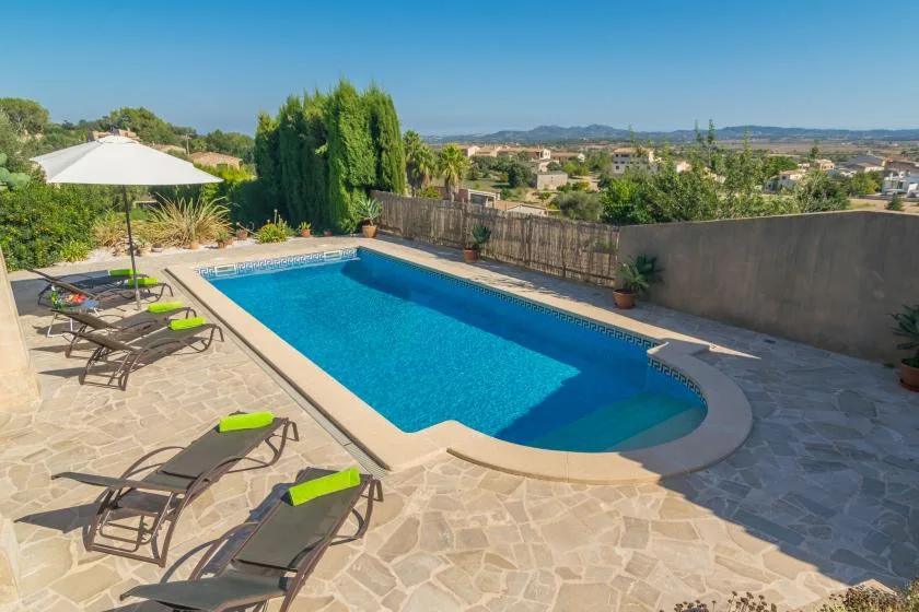 A swimming pool sits beside a stone patio with loungers and an umbrella.