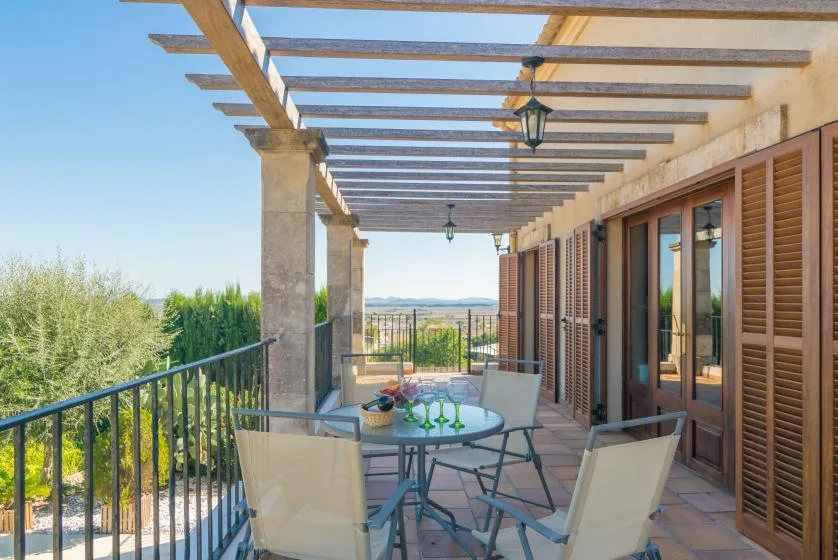A covered outdoor terrace has a table and seating beside shuttered doors.