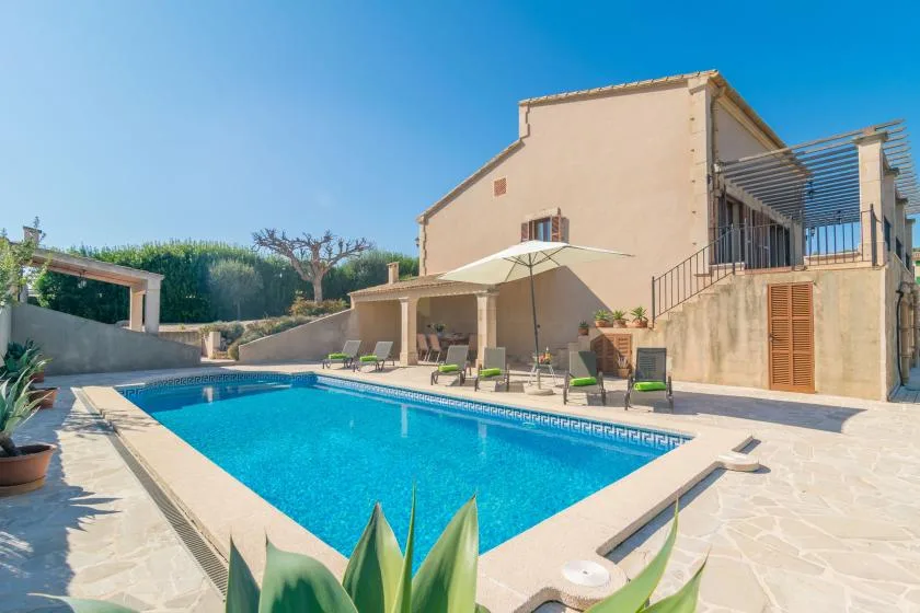 A swimming pool sits beside a tan house, with patio seating and an umbrella.