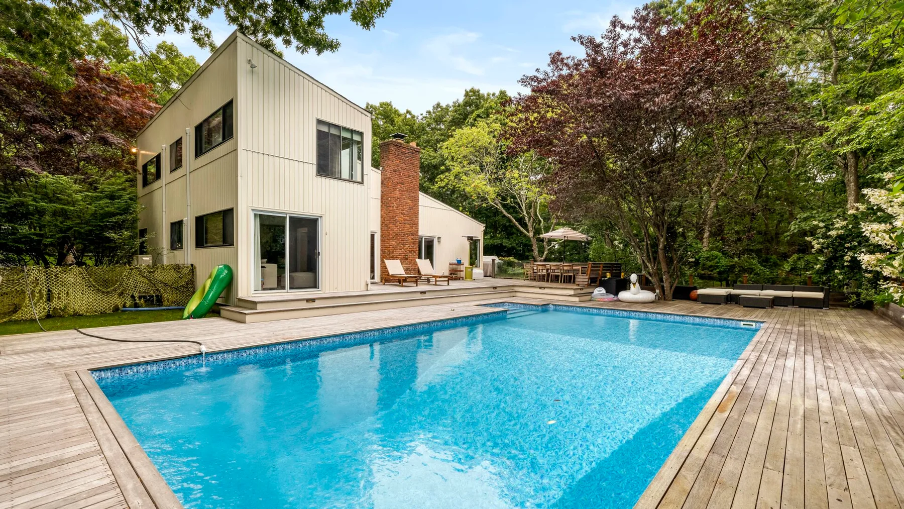 Una piscina y una terraza de madera están junto a una casa moderna rodeada de árboles.