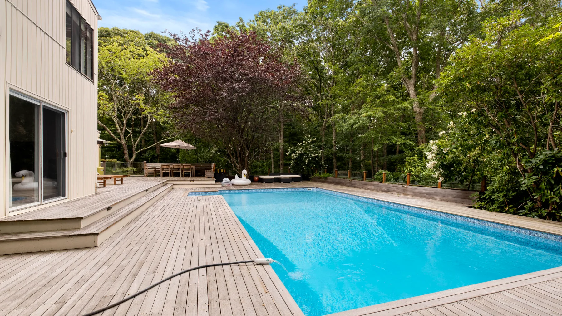 Una piscina al aire libre está junto a una terraza de madera y un jardín arbolado.