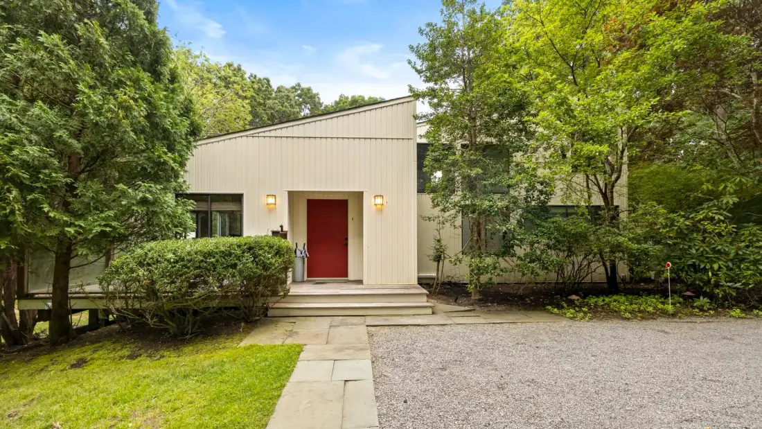 La entrada de una casa moderna con puerta roja, rodeada de árboles y arbustos.