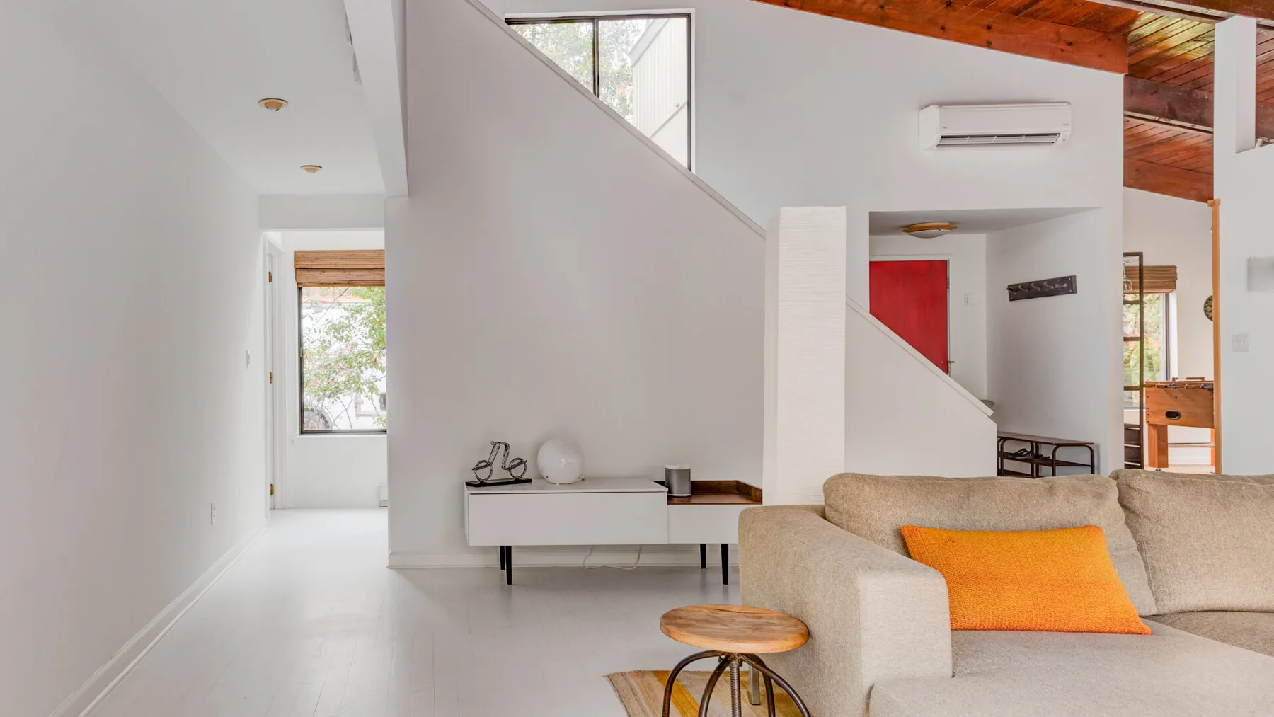 Una sala de estar luminosa con un sofá beige y un cojín naranja bajo una escalera.
