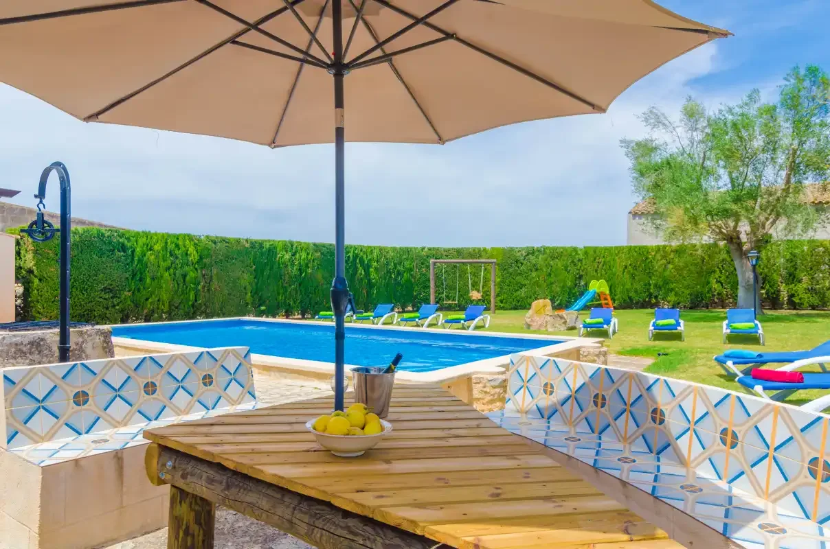Una piscina y tumbonas junto a un césped, con una mesa a la sombra en primer plano.