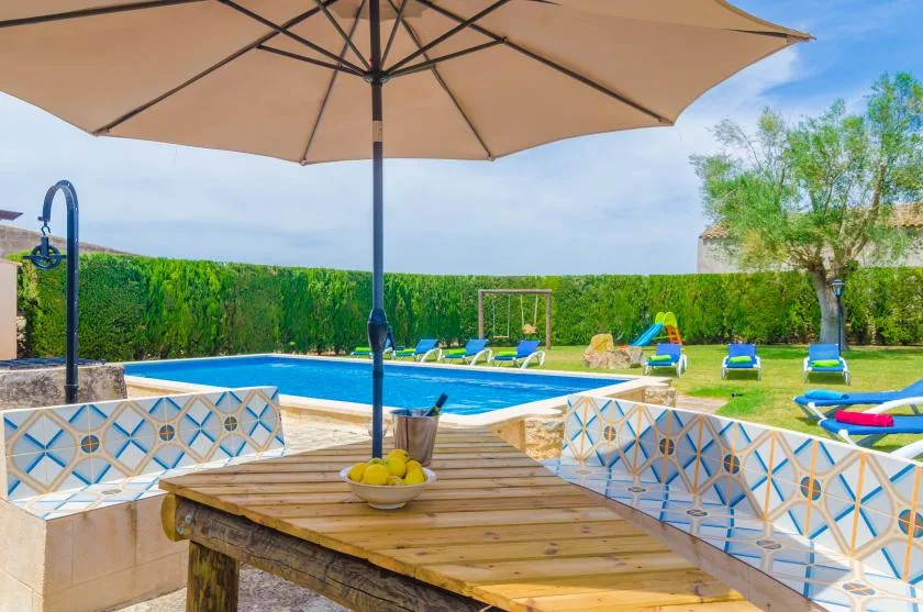 Una piscina al aire libre junto a una mesa de patio a la sombra y un césped verde.