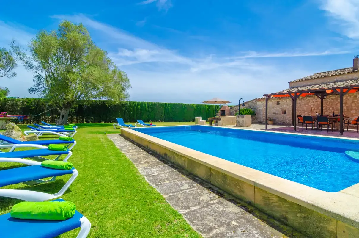 Una piscina junto a un césped y una zona de asientos a la sombra.