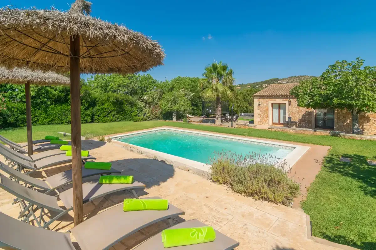 Una piscina exterior junto a un edificio de piedra, con tumbonas bajo una sombrilla de paja.