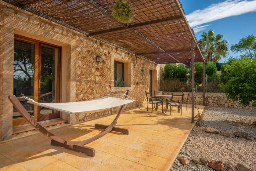 Patio exterior sombreado con una hamaca y una mesa de comedor junto a un muro de piedra.