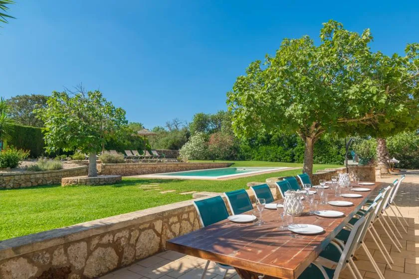 Una zona de comedor al aire libre con una mesa larga junto a una piscina y un césped verde.
