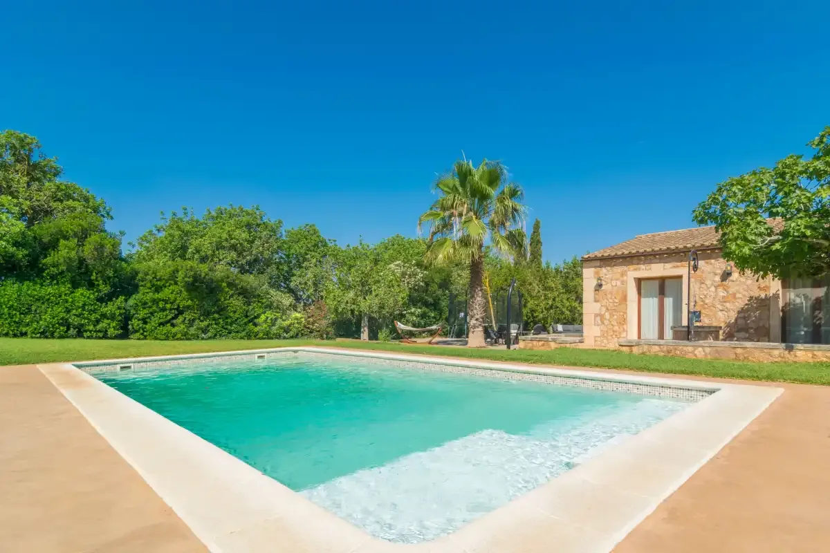 Una piscina junto a un edificio de piedra, con árboles y una palmera al fondo.