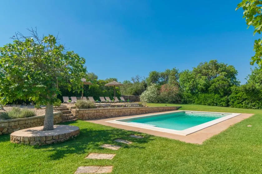 Una piscina al aire libre está junto a un césped y una zona de descanso con sombra.
