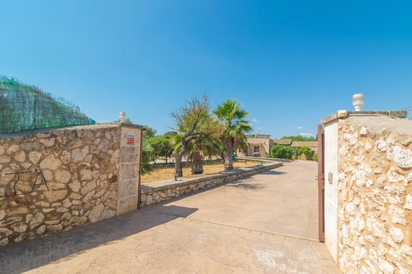Una entrada pavimentada se abre entre muros de piedra hacia un patio con palmeras.