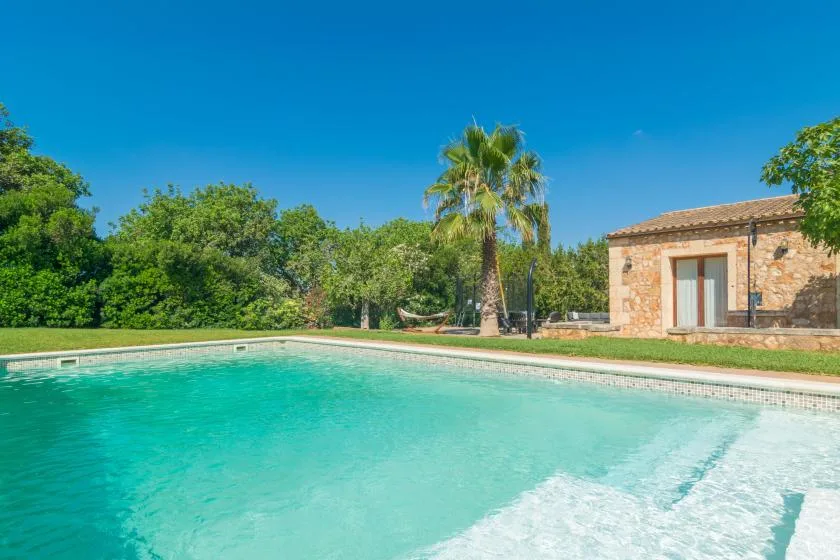 Una piscina junto a un edificio de piedra, con árboles y una palmera al fondo.