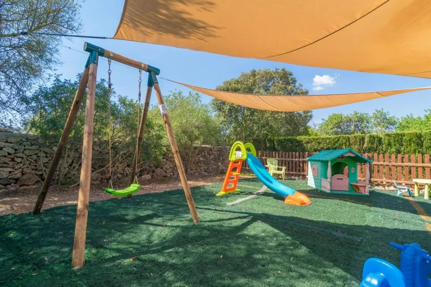 Una zona de juegos al aire libre con sombra tiene un columpio, un tobogán y una casita.