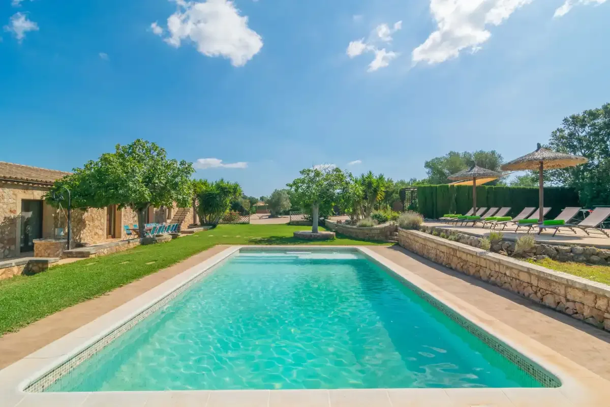 Una piscina está rodeada de césped, árboles y asientos a la sombra.