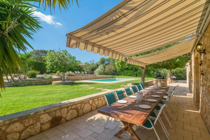 Una zona de comedor al aire libre con sombra da a un césped y una piscina.