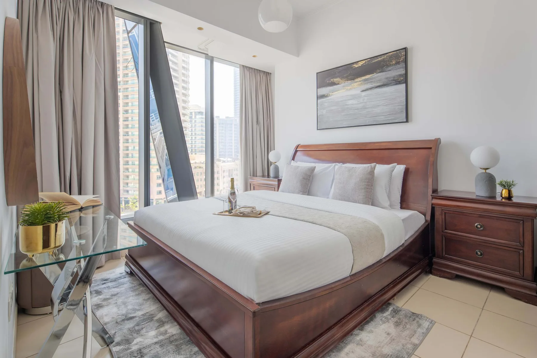 A bedroom with a large bed beside tall windows overlooking nearby buildings.