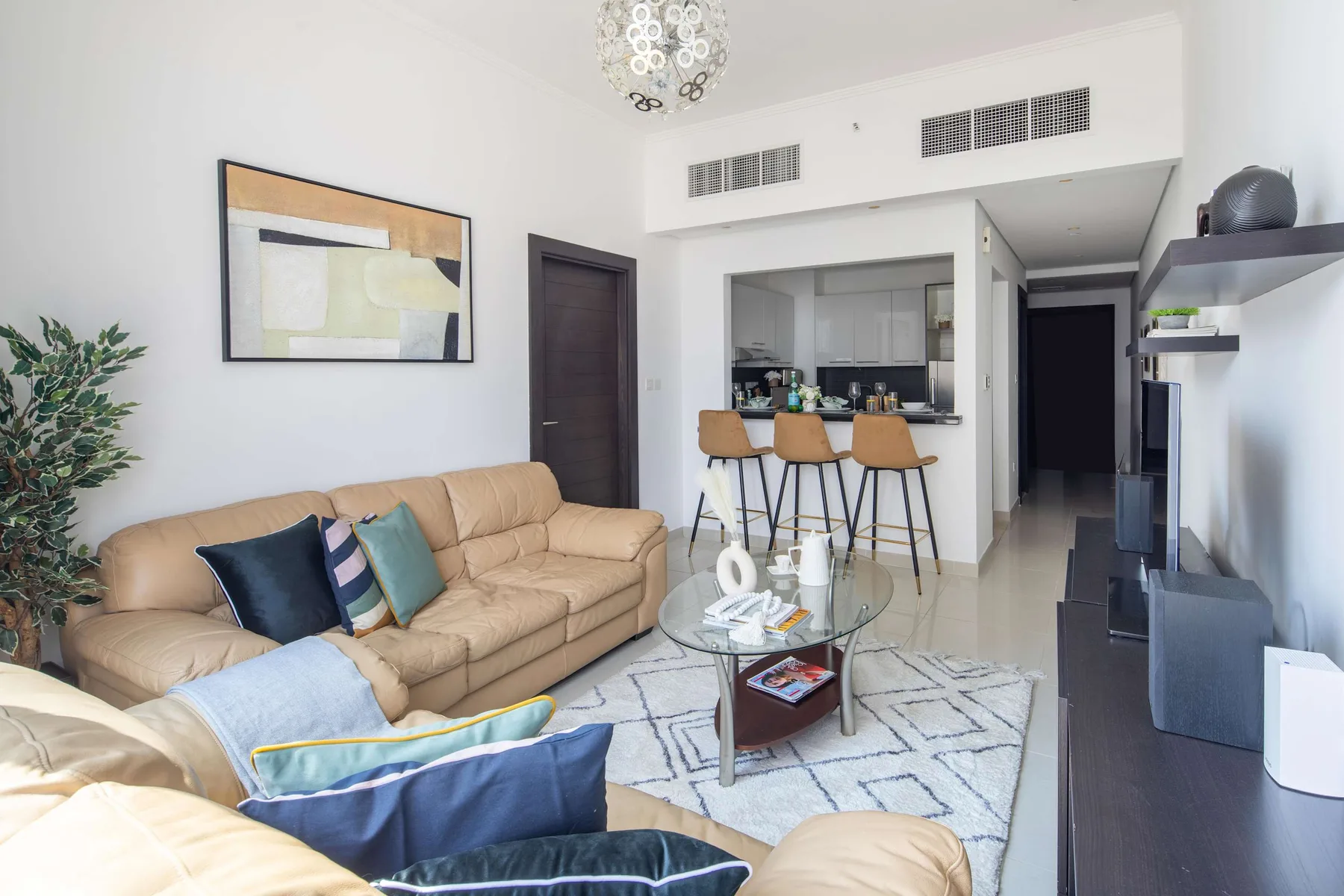 A bright living room with a beige sofa, glass coffee table, and open kitchen.