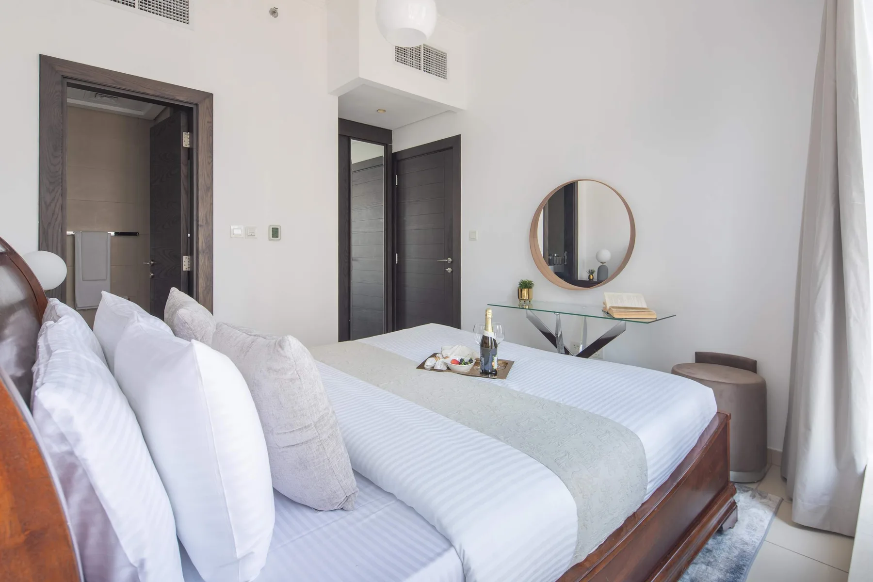 A bright bedroom with a neatly made bed and a round mirror above a desk.