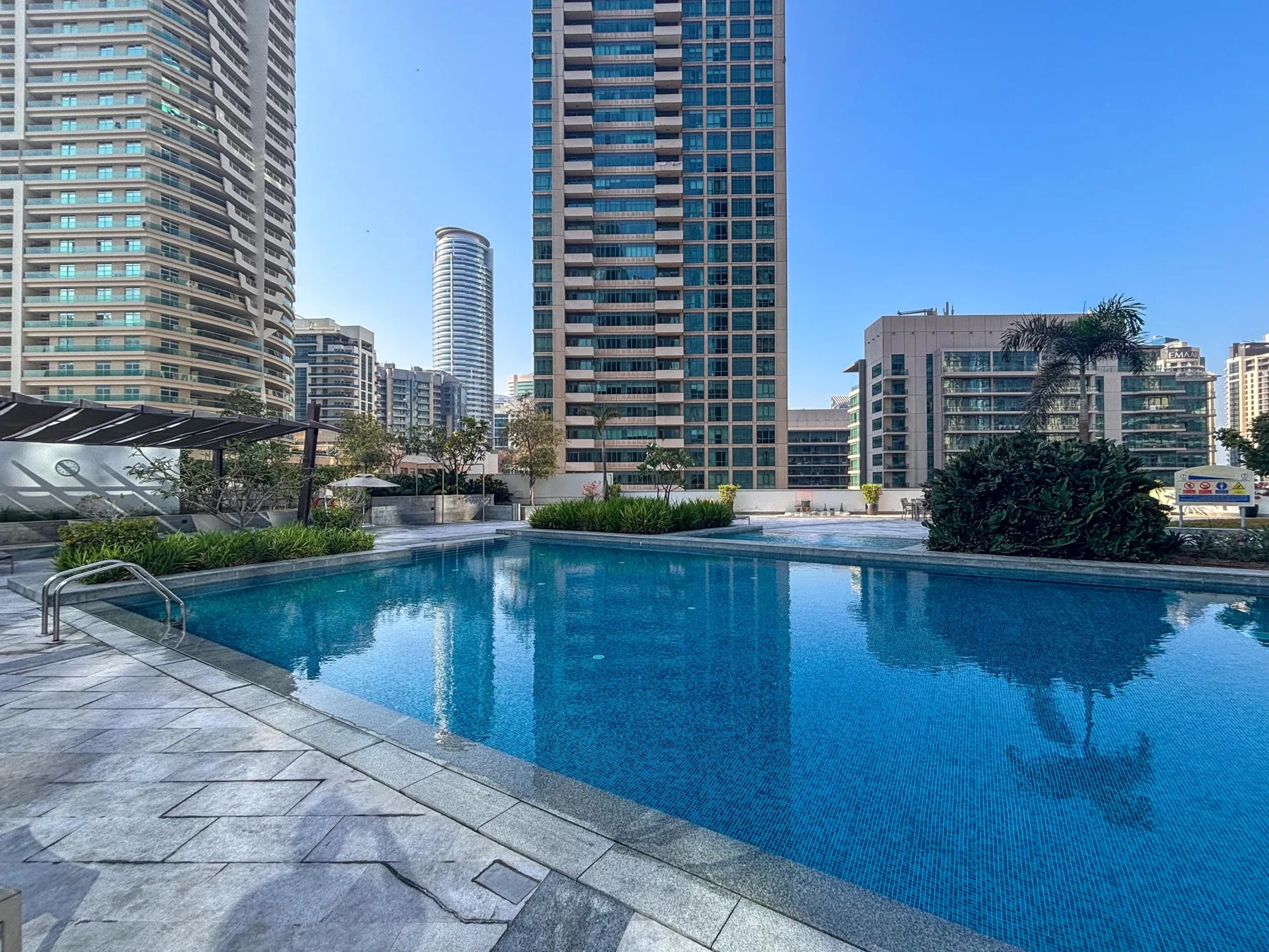 A swimming pool sits among tall buildings, with paved decking along the water.