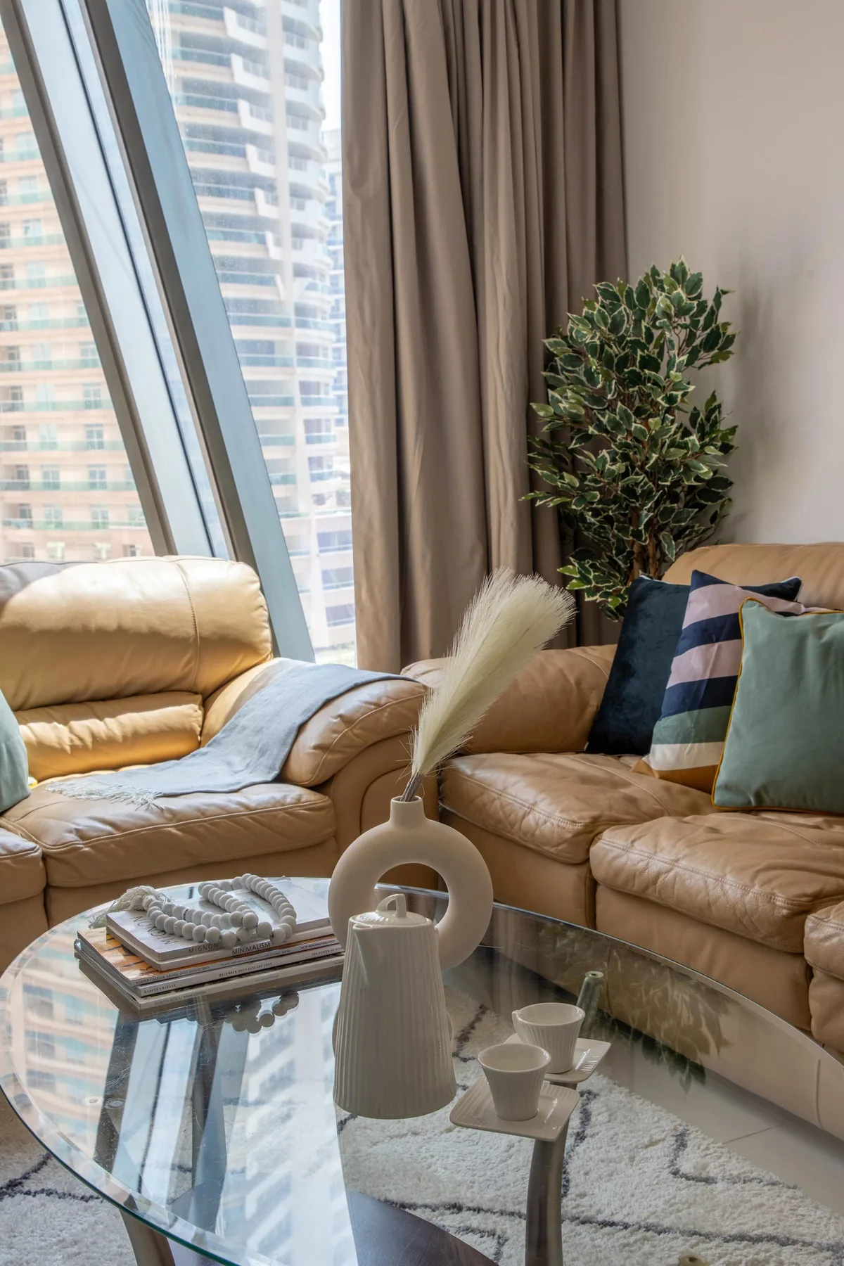 A living room with tan seating, a glass coffee table, and tall windows.